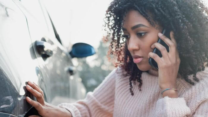Young lady of African background talking on the phone and looking at damage to her car