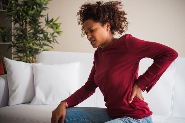 how do I know if my back pain is serious - black lady in red blouse sitting on the bed holding to her back and grimacing