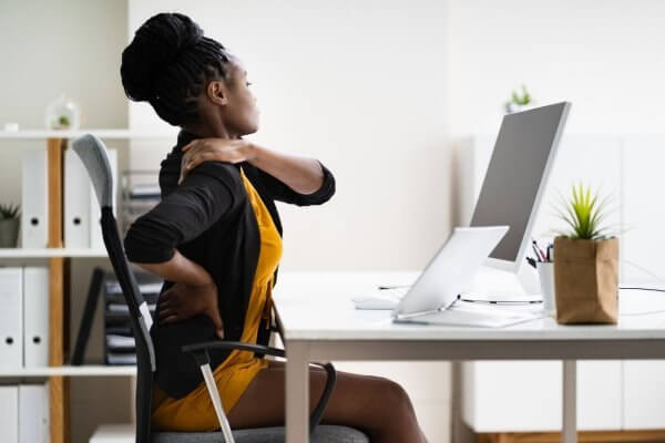 young african lady in gold dress and jacked seating at a workstation with serious back pain in her upper back