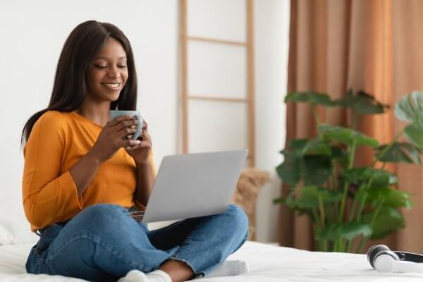 Black skinned lady sitting on her bed, looking at her laptop while sipping a cup of detox tea